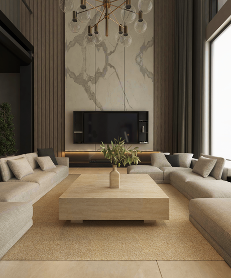 A modern living room with beige sectional sofas, a large light wood coffee table, a vase with greenery, a wall-mounted TV, marble wall accents, and a gold chandelier. Light streams in from a large window.
