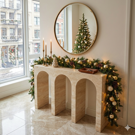 A modern entryway with a beige stone console table featuring three arches, two matching lamps with beige shades, a glass vase with green fern leaves, books, and a round mirror on the wall.