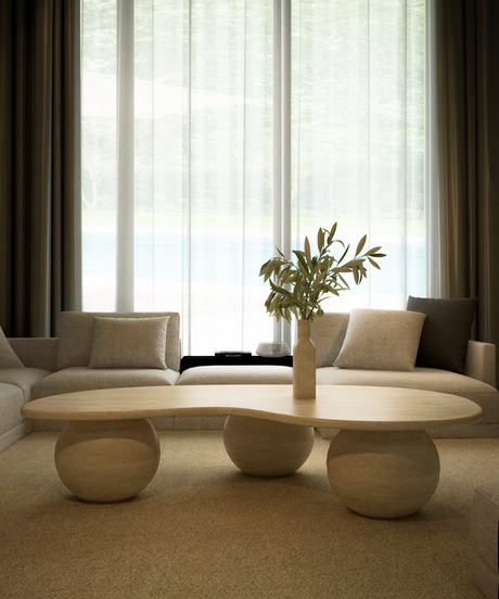 A modern living room with a light beige sofa, a sculptural coffee table with rounded legs, and a vase with green foliage, set in front of tall windows with sheer curtains.