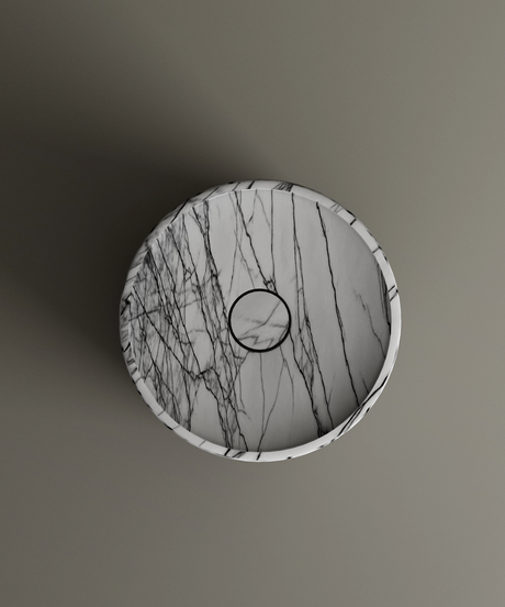 A round, white marble sink with gray and black veining is shown from above against a plain, taupe-colored background. The sink is empty and the drain is visible at the center.