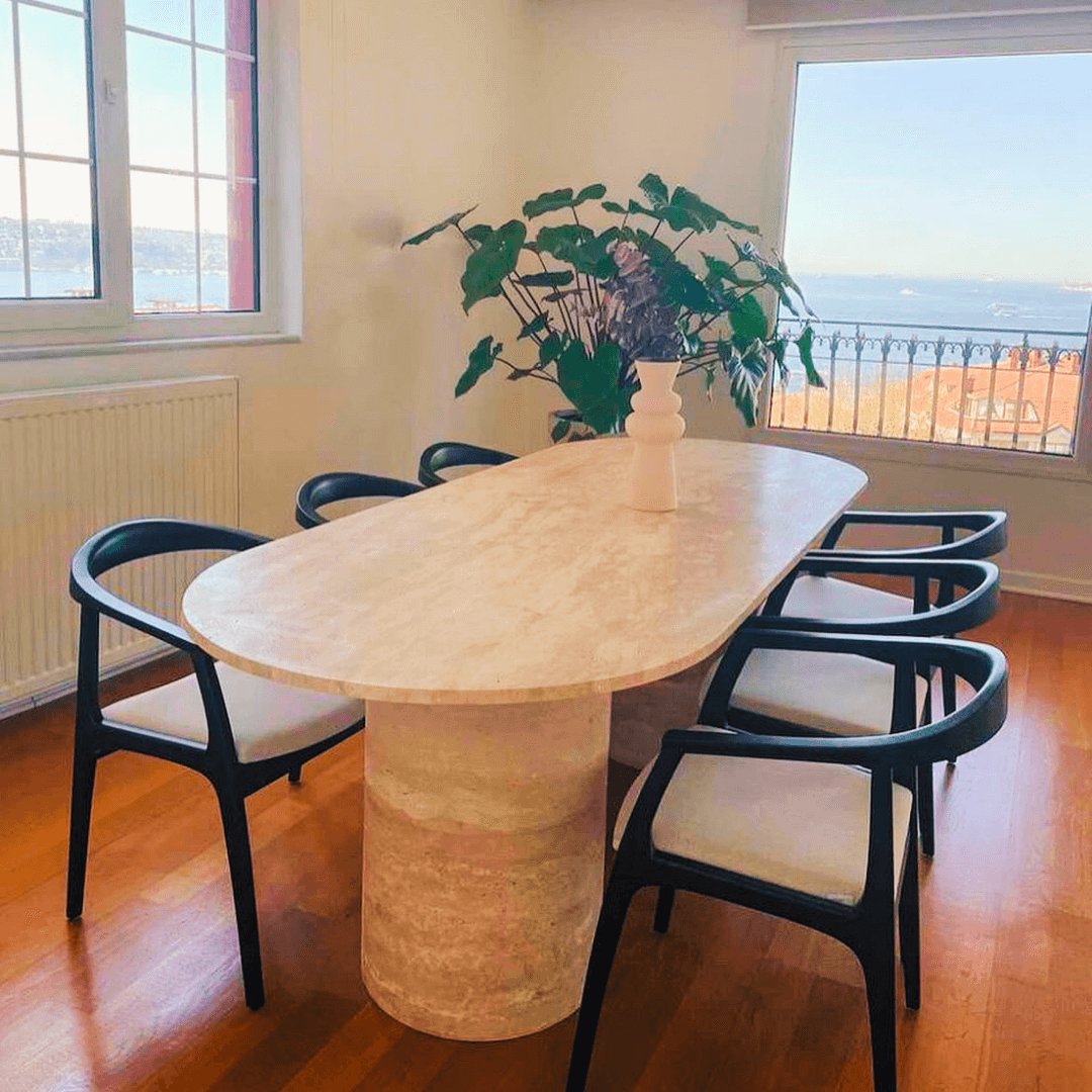 The dining room features an Arcadia Dining Table by Travertable, showcasing an elegant oval design. Surrounded by six black chairs with white cushions, a white vase with green plants adds charm. Large windows offer stunning sea views and distant hills, while a warm wooden floor enhances elegance.