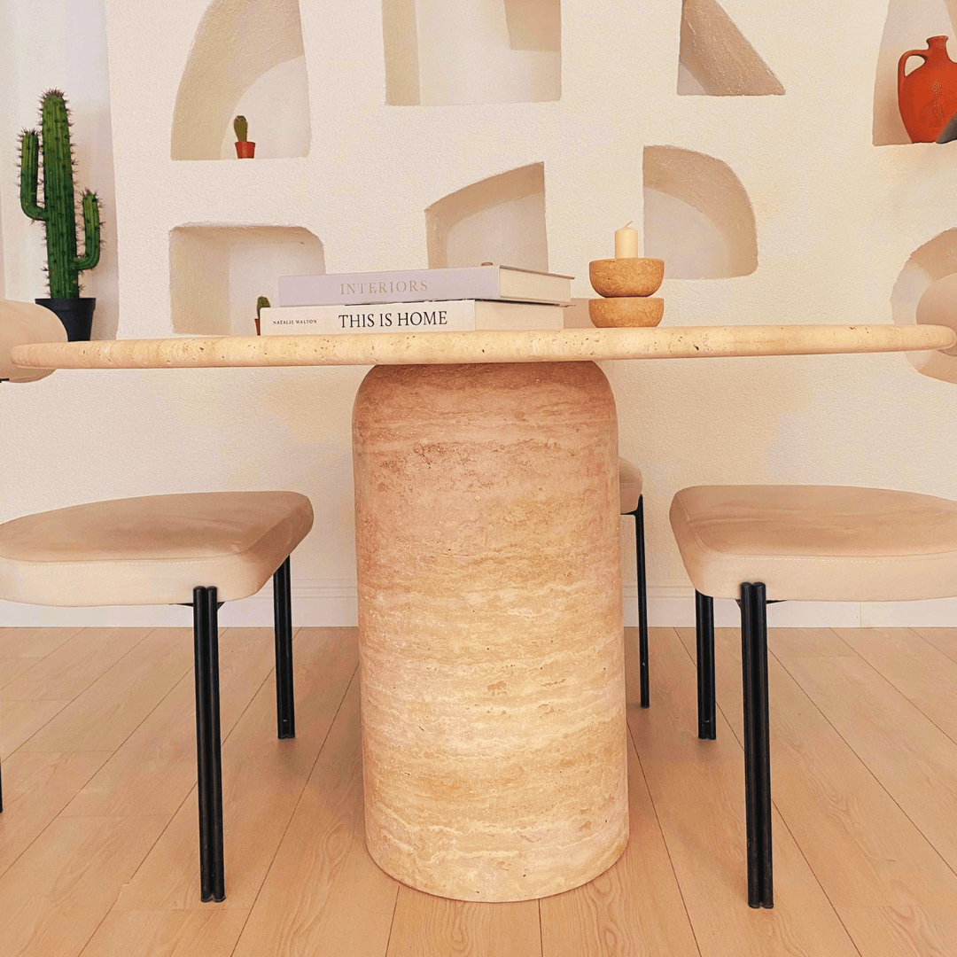 A modern dining area showcases Travertables Chic Oval Travertine Dining Table accompanied by beige cushioned chairs. Books, a candle, and a small bowl adorn the tabletop. A cactus and decorative wall niches enhance the setting, all beautifully laid out on a light wooden floor.