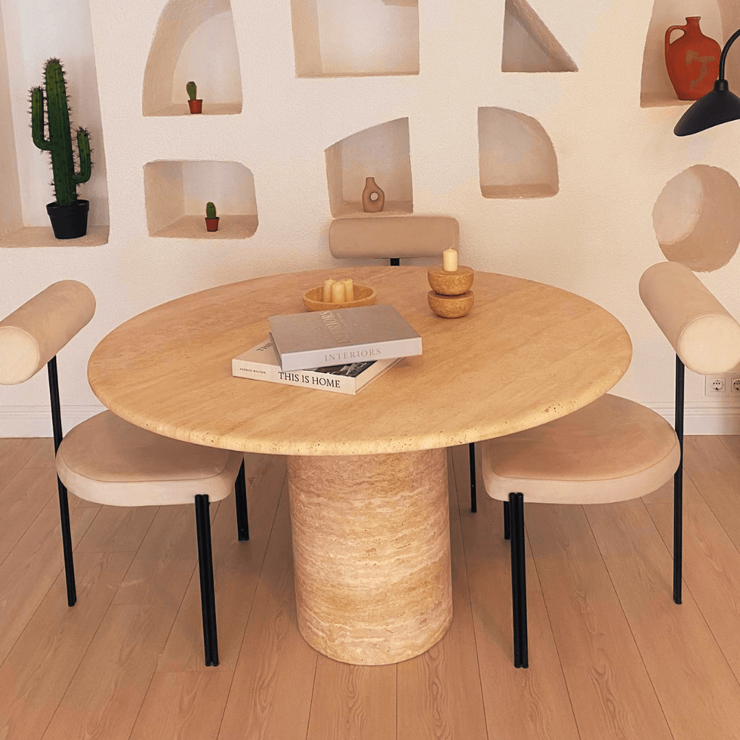 The Chic Oval Travertine Dining Table by Travertable, with a central cylindrical base, is encircled by four beige chairs featuring black legs. Books, candles, and bowls adorn the table while recessed white shelves in the background display a small cactus and decor items.