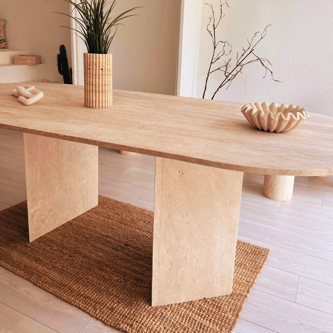 A minimalist living room showcases The Grand Pillar Travertine Dining and Meeting Table by Travertable on a woven rug, adorned with a potted plant and ceramic decor. Bare branches in a vase are highlighted by soft natural light, enhancing the serene atmosphere of the modern design.