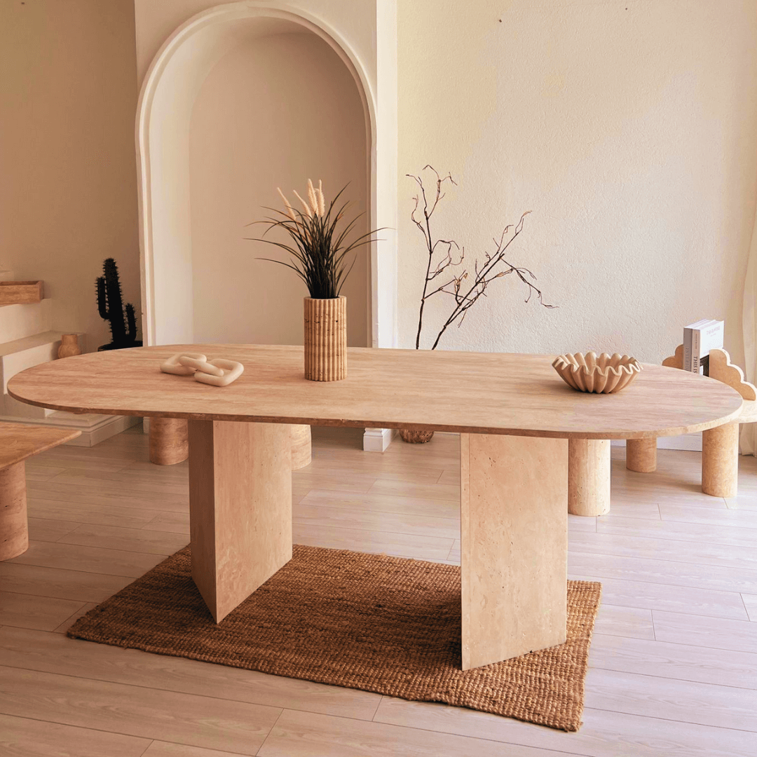 In a modern room, the Travertable Grand Pillar Travertine Dining and Meeting Table with customizable wooden oval top and geometric legs sits on a woven rug. Displaying a tall vase of pampas grass and a wavy ceramic bowl, it complements the premium travertine backdrop featuring beige arches.