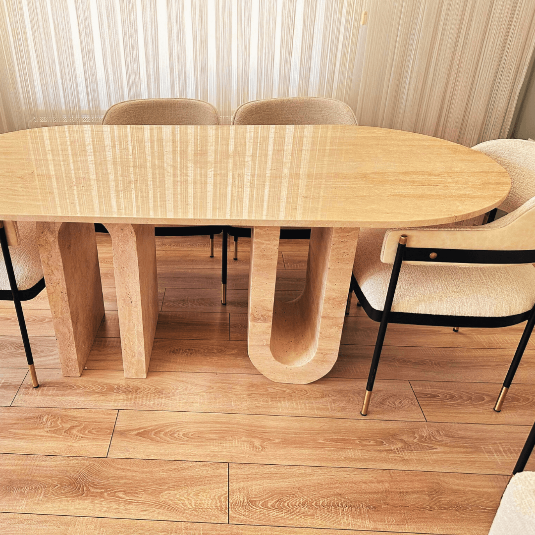 The Travertable Luxurious Oval Travertine Dining Table with Dual Columns is paired with upholstered chairs on a wooden floor. The room radiates warmth, enhanced by sunlight streaming through sheer curtains, creating an inviting and cozy atmosphere.