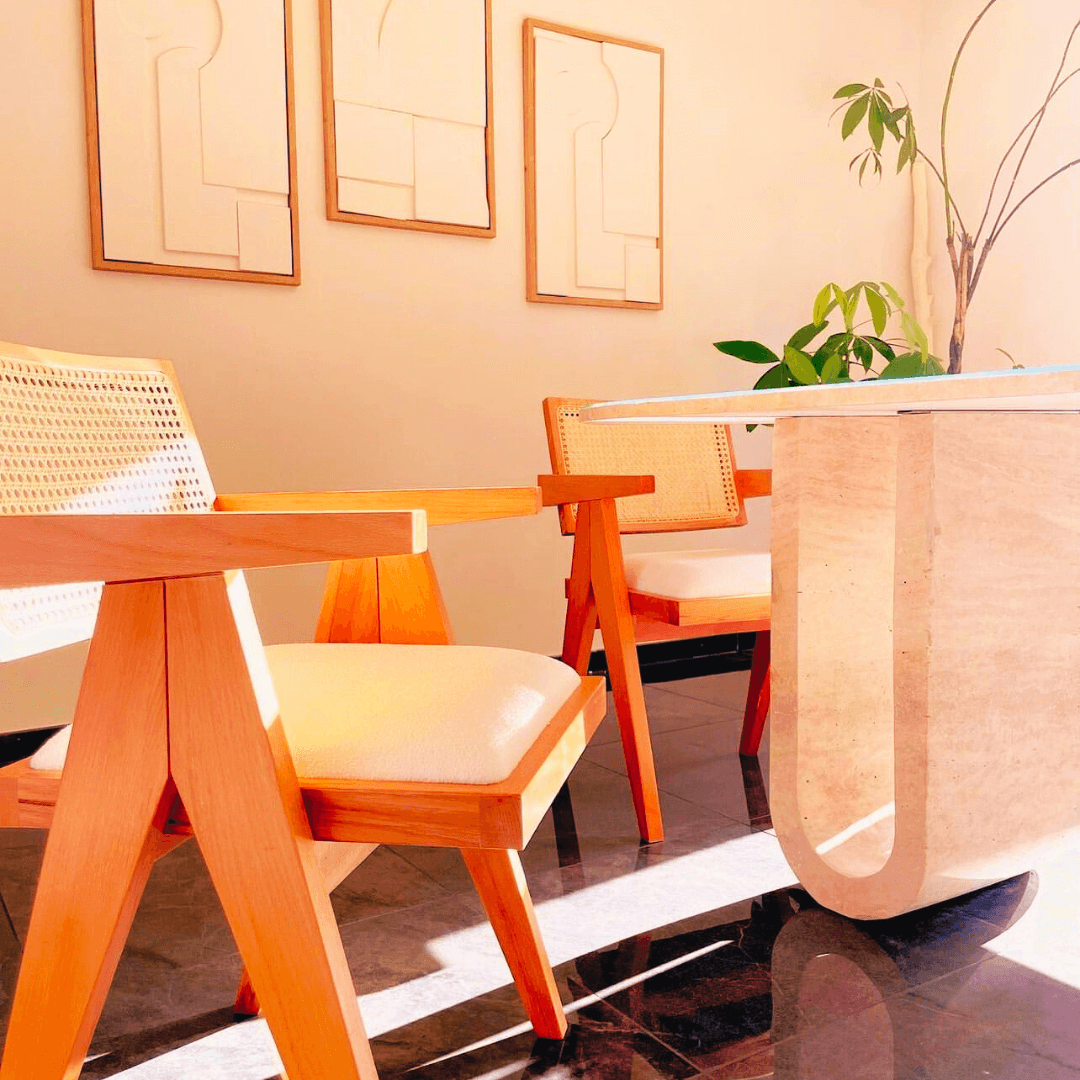 A sunlit room showcases a modern wooden desk and two wooden armchairs with woven seats. Three abstract art pieces adorn the wall, complemented by a small plant for greenery. The space is anchored by Travertables Luxurious Oval Travertine Dining Table with Dual Columns, adding an elegant and inviting touch.