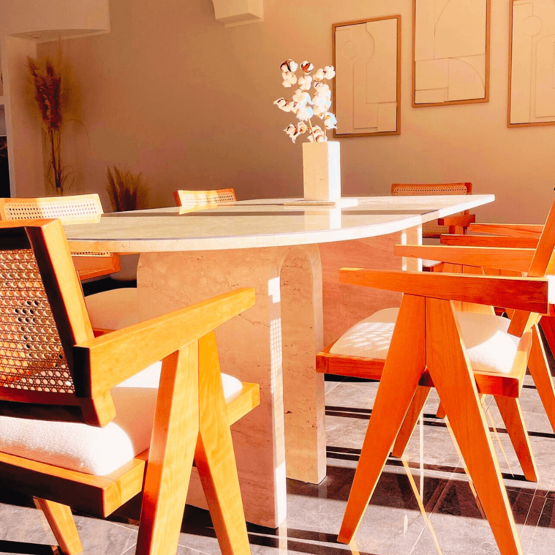 A modern dining room features a Luxurious Oval Travertine Dining Table with Dual Columns by Travertable, encircled by wooden chairs with white cushions. A vase of white flowers and minimalist art decorate the warm beige walls as sunlight creates a cozy ambiance in this elegant space.
