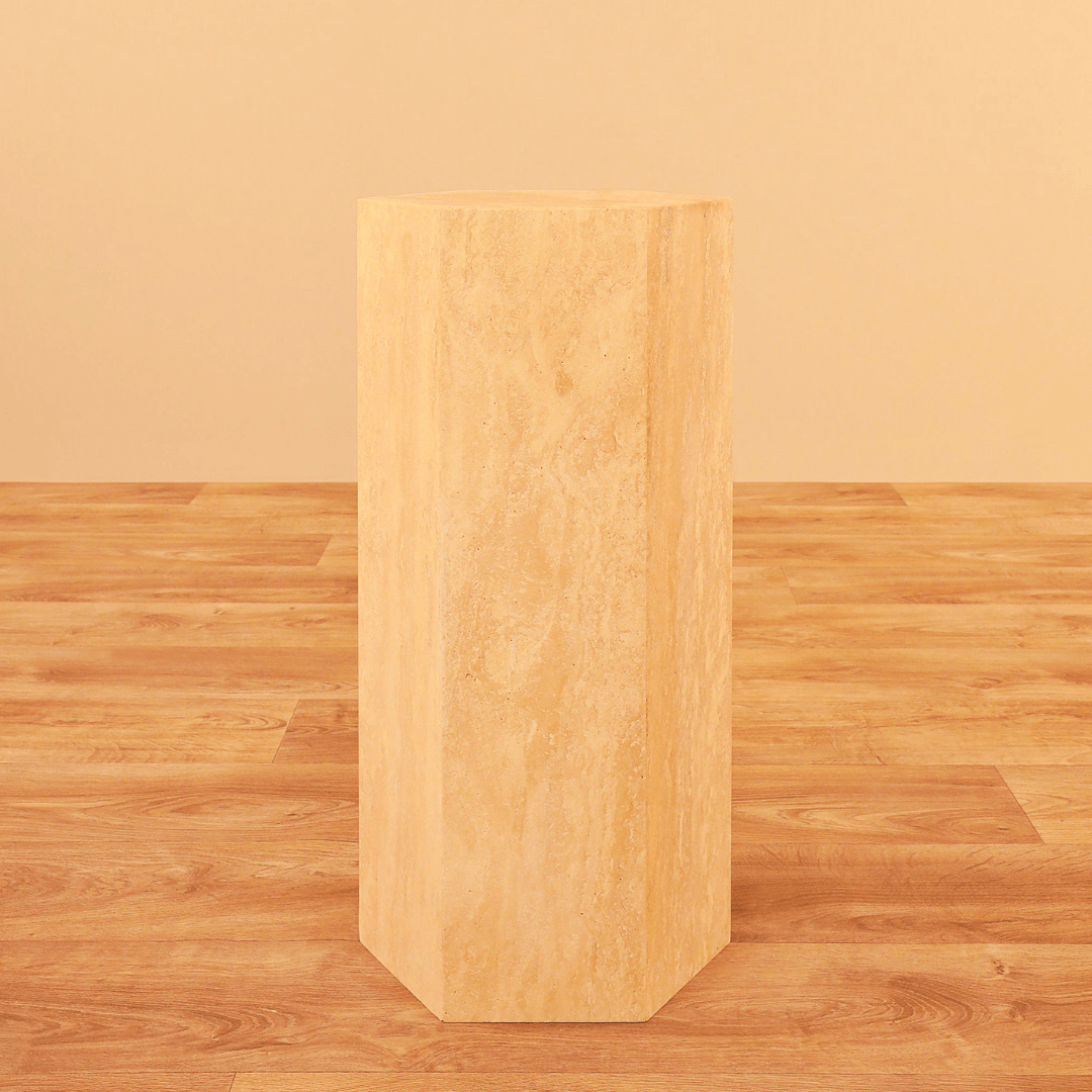 The Geometric Travertine Pedestal by Travertable, featuring a unique sculptural design and architectural appeal, stands tall like a hexagonal column on a matching wooden floor against a beige background.