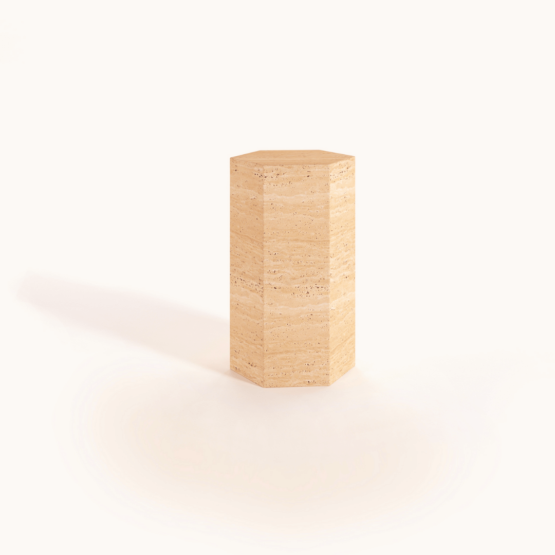 The Geometric Travertine Pedestal by Travertable is a tall, hexagonal column crafted from light brown speckled stone. It stands against a white background, with its polished surface reflecting light to emphasize its unique sculptural design and architectural appeal.