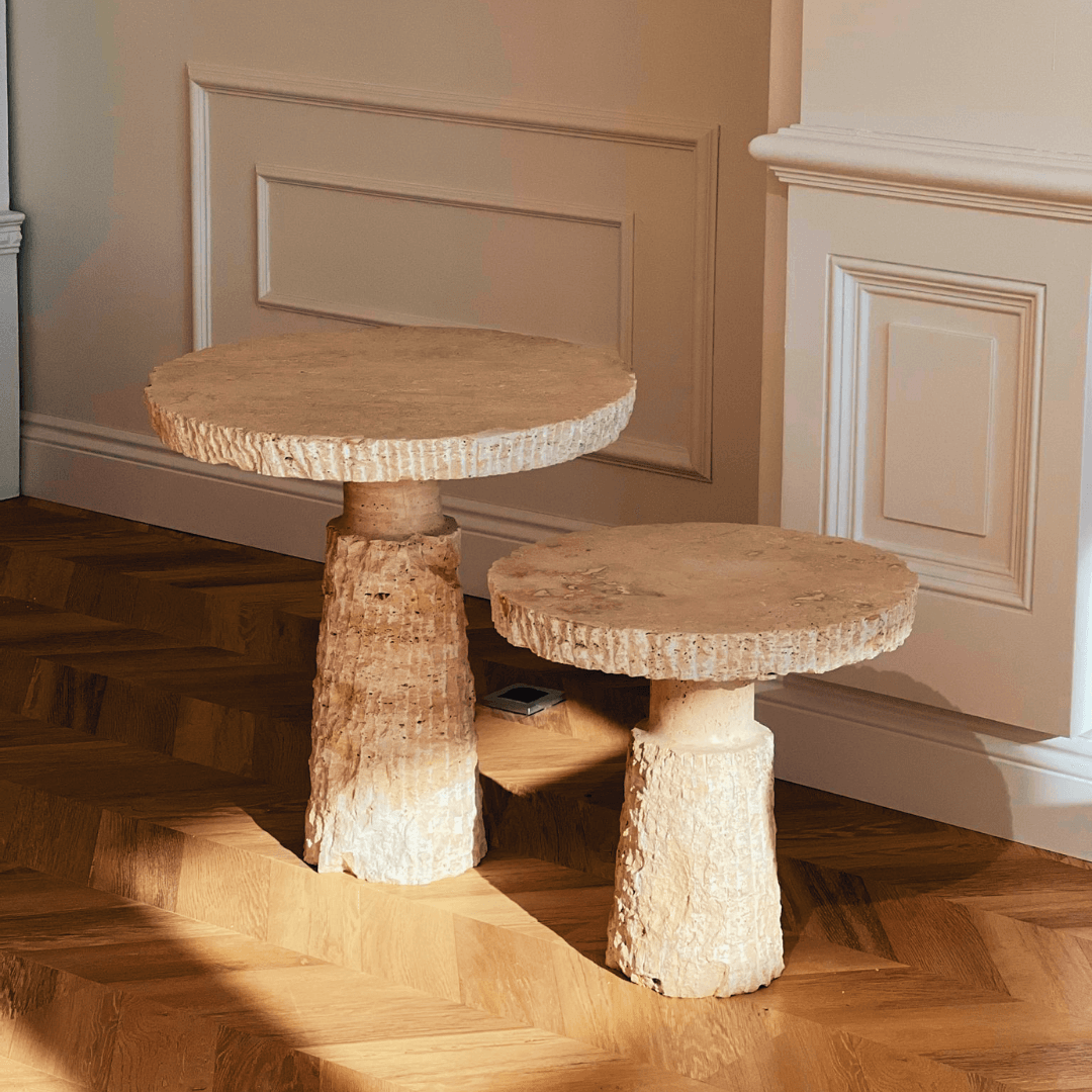 Two Handcrafted Travertine Marble Side Tables by Travertable with textured surfaces rest on a herringbone wood floor. Sunlight casts soft shadows, enhancing their elegant look against cream-colored paneled walls.