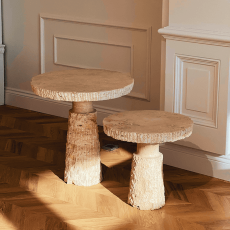 Two Handcrafted Travertine Marble Side Tables by Travertable with textured surfaces rest on a herringbone wood floor. Sunlight casts soft shadows, enhancing their elegant look against cream-colored paneled walls.