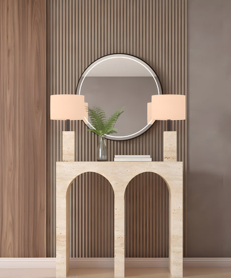 The TRAVERTABLE Travertine Console Table with Two Arches, crafted from solid stone, holds matching lamps, a vase of green ferns, and a closed book. A round mirror on the paneled brown wall completes this modern, sophisticated look.