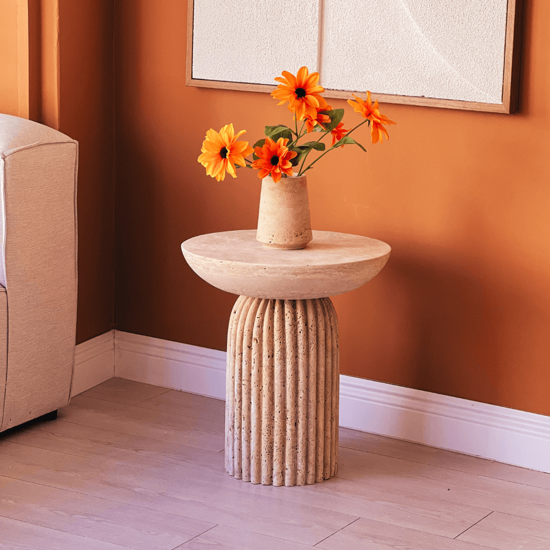 A Fluted Travertine Side Table with a sculptural column base by Travertable, sits in an orange-walled room, adorned with a matching vase and vibrant orange flowers. Part of a beige sofa is visible on the left.