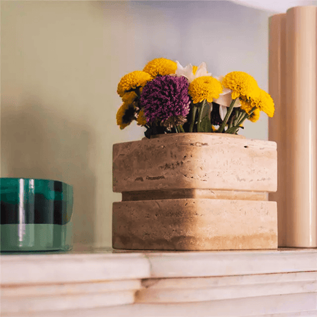A Travertable Vintage Charm Travertine Vase holds yellow and purple flowers on a marble surface. Nearby is a green and black striped container, with a tall beige cylindrical object in the background against a light wall.