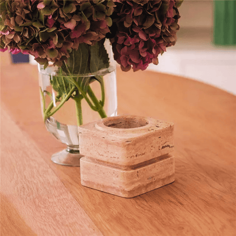 A Vintage Charm Travertine Vase by Travertable, filled with pink and purple hydrangeas, sits alongside a square stone candle holder on a wooden table. The setting offers a warm and cozy ambiance with an emphasis on natural materials.