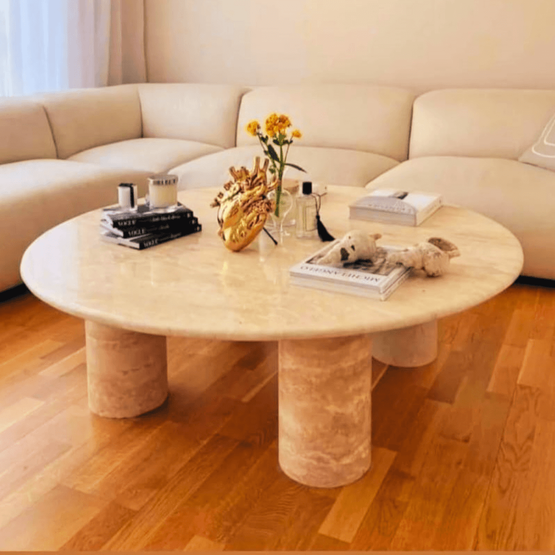 The Natural Retreat Travertine Coffee Table by Travertable, featuring a round design with cylindrical legs, adorns the wooden floor and holds books, a vase of yellow flowers, decor accents, and stuffed animals. In the background is a plush beige sectional sofa framed by sheer curtains—luxury epitomized.