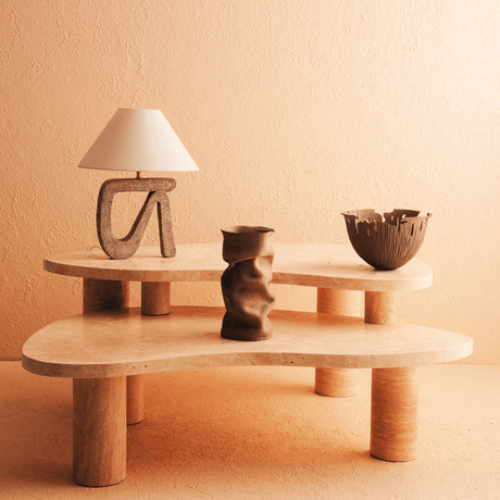 A Vintage-Inspired Travertable coffee table with unique organic veining, two irregular tiers, and textured legs sits against a peach-toned wall, adorned with a lamp featuring a curved base and conical shade, accompanied by a textured vase and bowl.