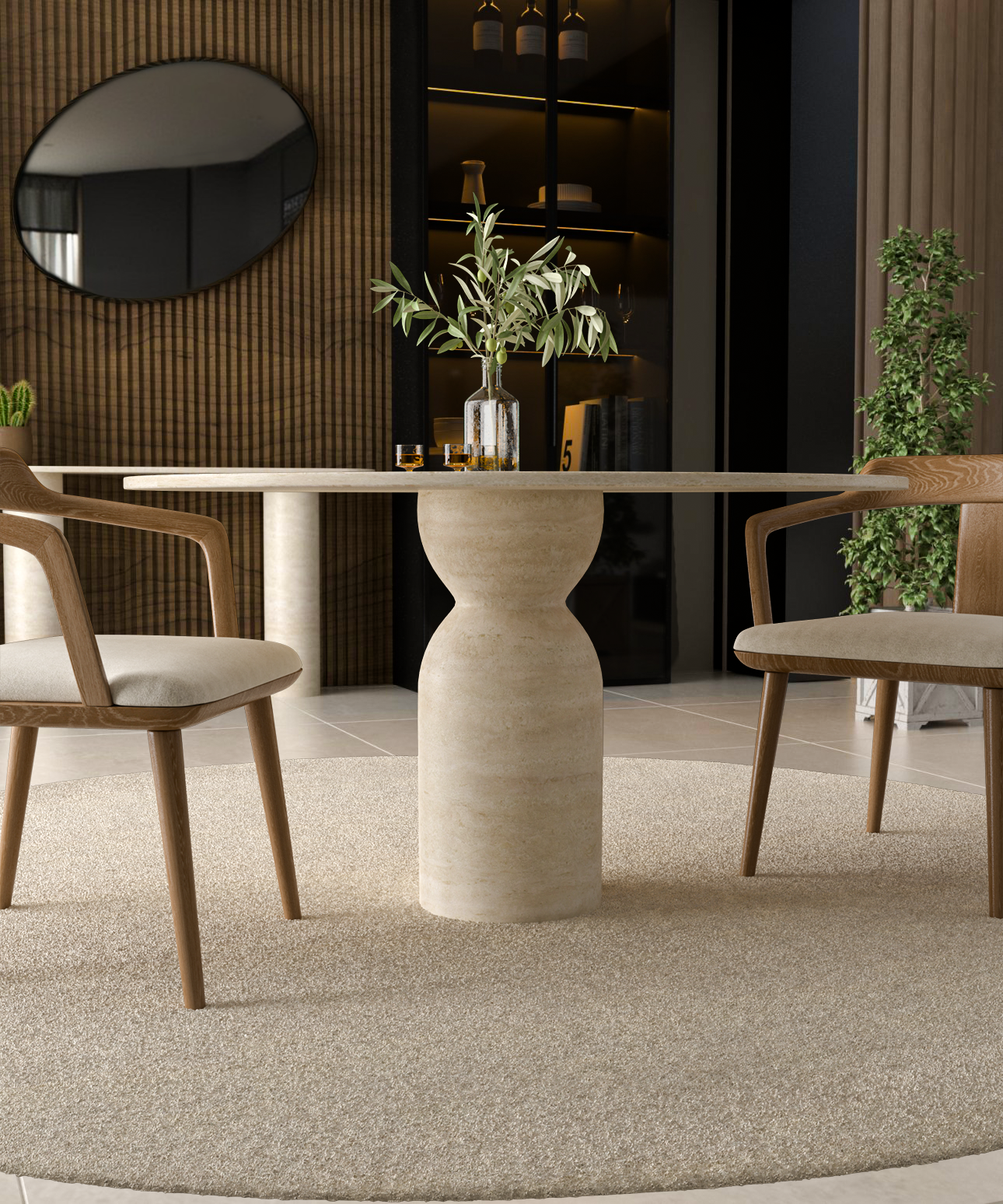 The TRAVERTABLE Oval Travertine Dining Table with a sculptural round center base stands on a beige rug between two wooden chairs. A glass bottle with branches and two glasses sit on the table, set against a wall mirror, plants, and a wooden accent wall.