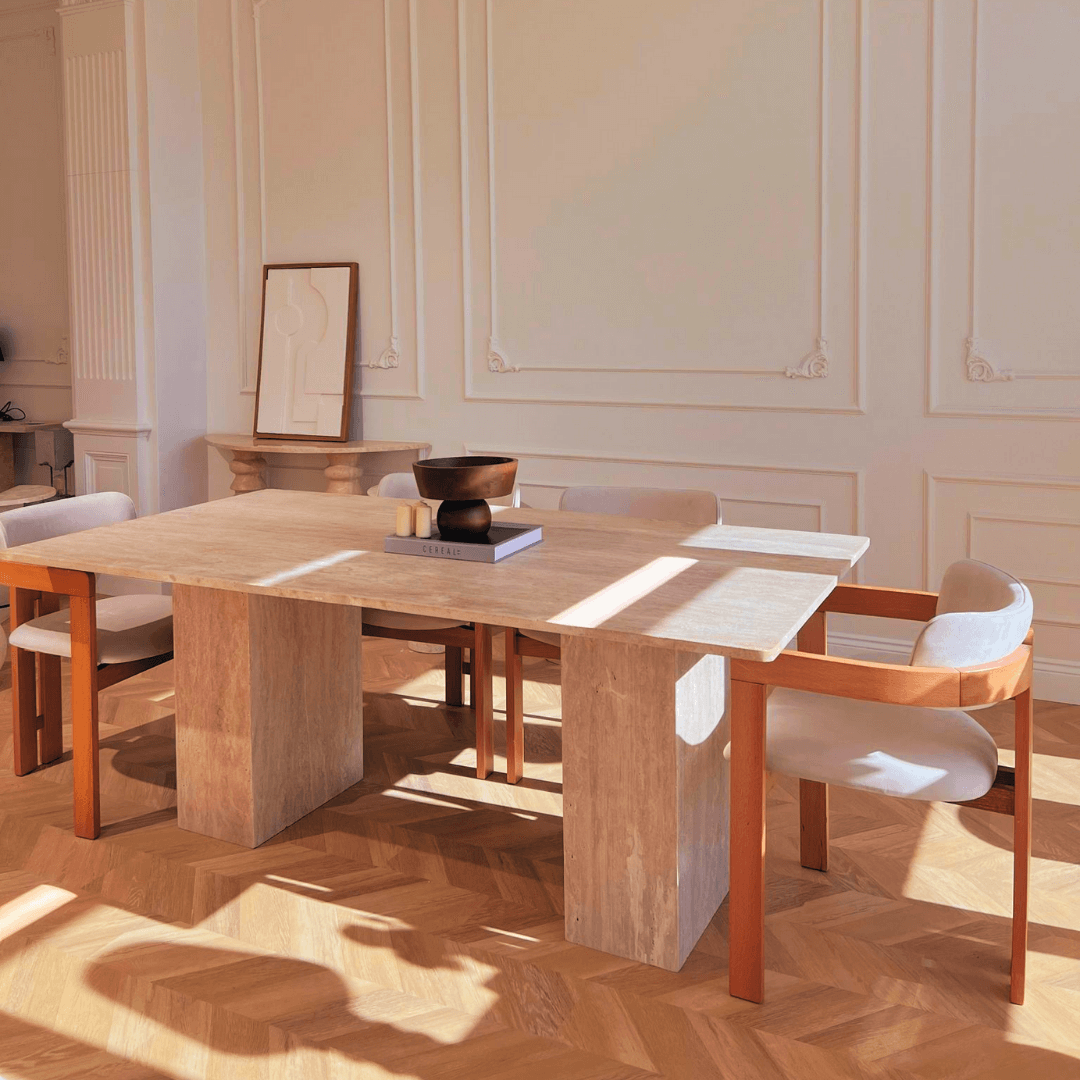 A sunlit dining room features a modern design with The Grand Travertine Dining Table by Travertable, surrounded by four cushioned chairs with wooden arms on a herringbone parquet floor. Minimalistic artwork leans against a paneled white wall, and a dark bowl sits elegantly on the table.