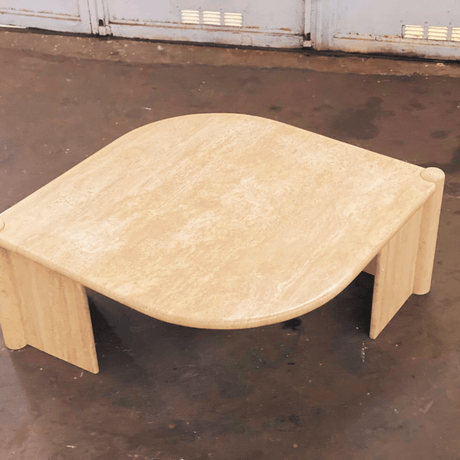 The Travertable Travertine Grace Coffee Table showcases natural beauty with its elegant Italian travertine and smooth polished surface, set on a dark floor. Its neutral beige tone and minimalist design contrast against light blue metal lockers in the background.