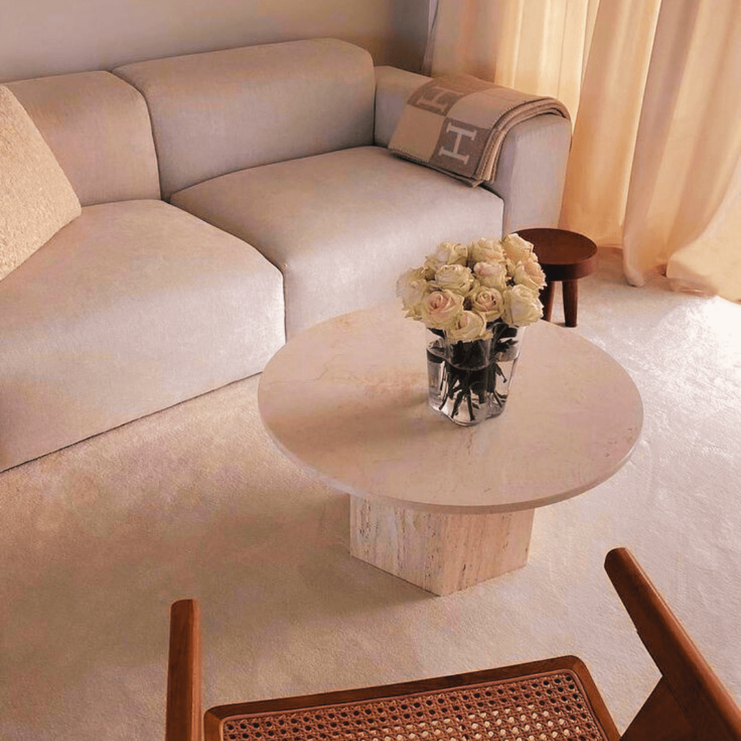 A cozy living room includes a light beige sofa and the Desert Serenity Travertine Coffee Table by Travertable, with a glass vase of white roses. A brown stool, tan throw blanket, and wooden chair with a woven seat complete the design as soft natural light filters through sheer curtains.