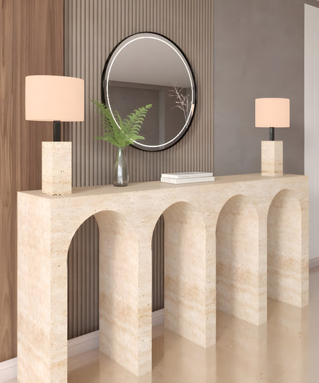 The TRAVERTABLE Travertine Console Table with Four Arches displays two square-based beige lamps, a glass vase with green fern leaves, and a stack of books. A round mirror hangs on the ribbed wall behind this architectural stone console.