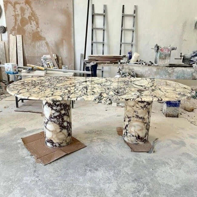 An Arcadia Dining Table by Travertable with a unique, intricate travertine pattern and two cylindrical bases stands in a workshop, surrounded by various tools, materials, and a ladder against the wall, indicating an active work environment.