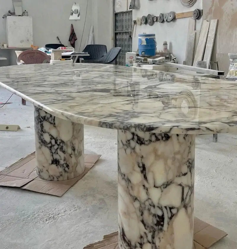 The Arcadia Dining Table by Travertable, with its polished marble rectangular top and two cylindrical pedestals in a black and white pattern, stands proudly in the workshop. Customize your table to suit any space with tools and chairs visible in the background.