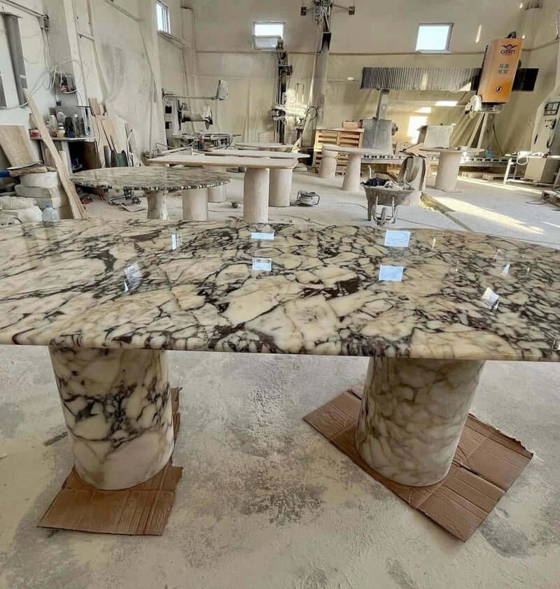 In a workshop, an Arcadia Dining Table by Travertable features a glossy, patterned marble surface on two cylindrical bases. It invites customization amid stonework materials and tools, alongside partially assembled travertine dining pieces.