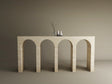 The TRAVERTABLE Travertine Console Table with Four Arches stands against a beige wall, highlighting sculptural elegance. Three minimalist decor pieces on top complete this refined display of the customizable Architectural Stone Console.