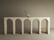 The TRAVERTABLE Travertine Console Table with Five Arches stands against a brown wall, topped with three white vases and a glass vase of bare branches—a customizable, sculptural masterpiece of understated luxury.