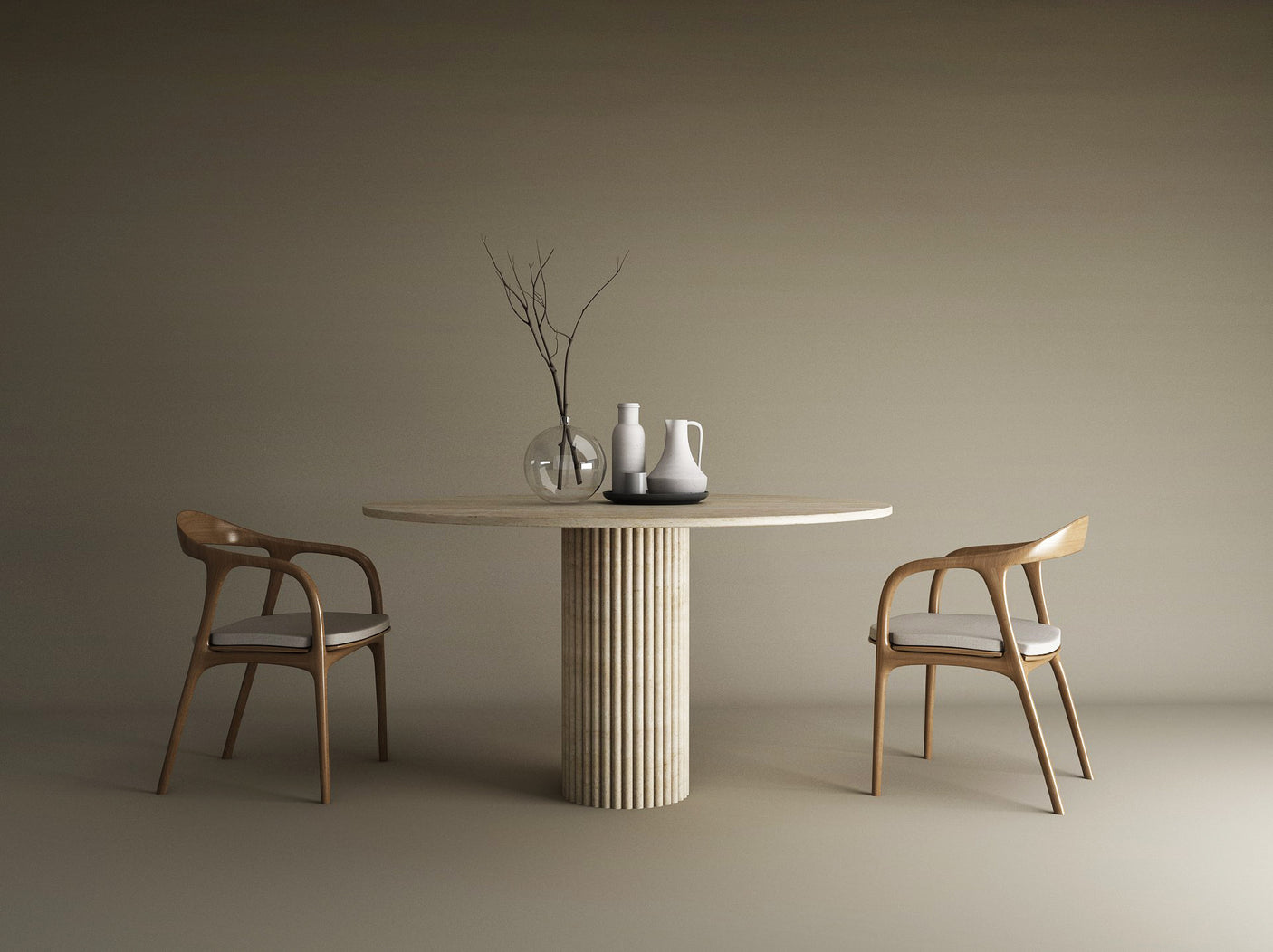 An Oval Travertine Dining Table with a fluted round base by TRAVERTABLE sits between two wooden chairs with white cushions. On the table are three minimalist vessels and a glass vase holding bare branches, set against a plain beige background.