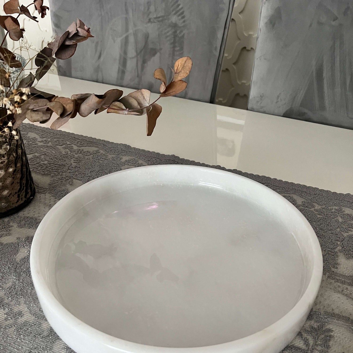 The Travertable White Alabaster Round Tray, a luxe marble accent piece, sits on a gray patterned table runner. To the left, a vase with dried branches adds to the minimalist elegance and luxury home decor of the tabletop arrangement.