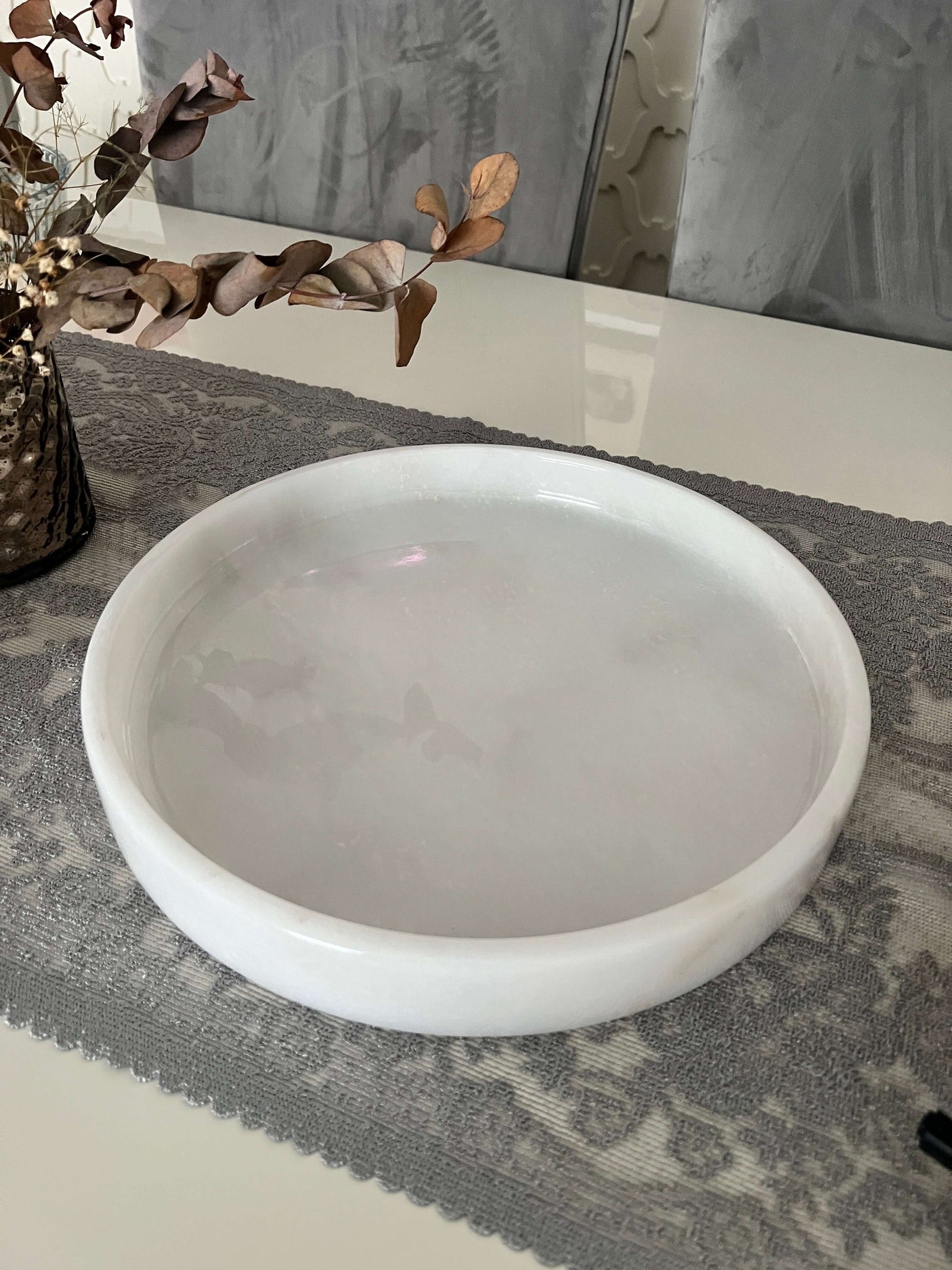 The Travertable White Alabaster Round Tray, a luxe marble accent piece, sits on a gray patterned table runner. To the left, a vase with dried branches adds to the minimalist elegance and luxury home decor of the tabletop arrangement.