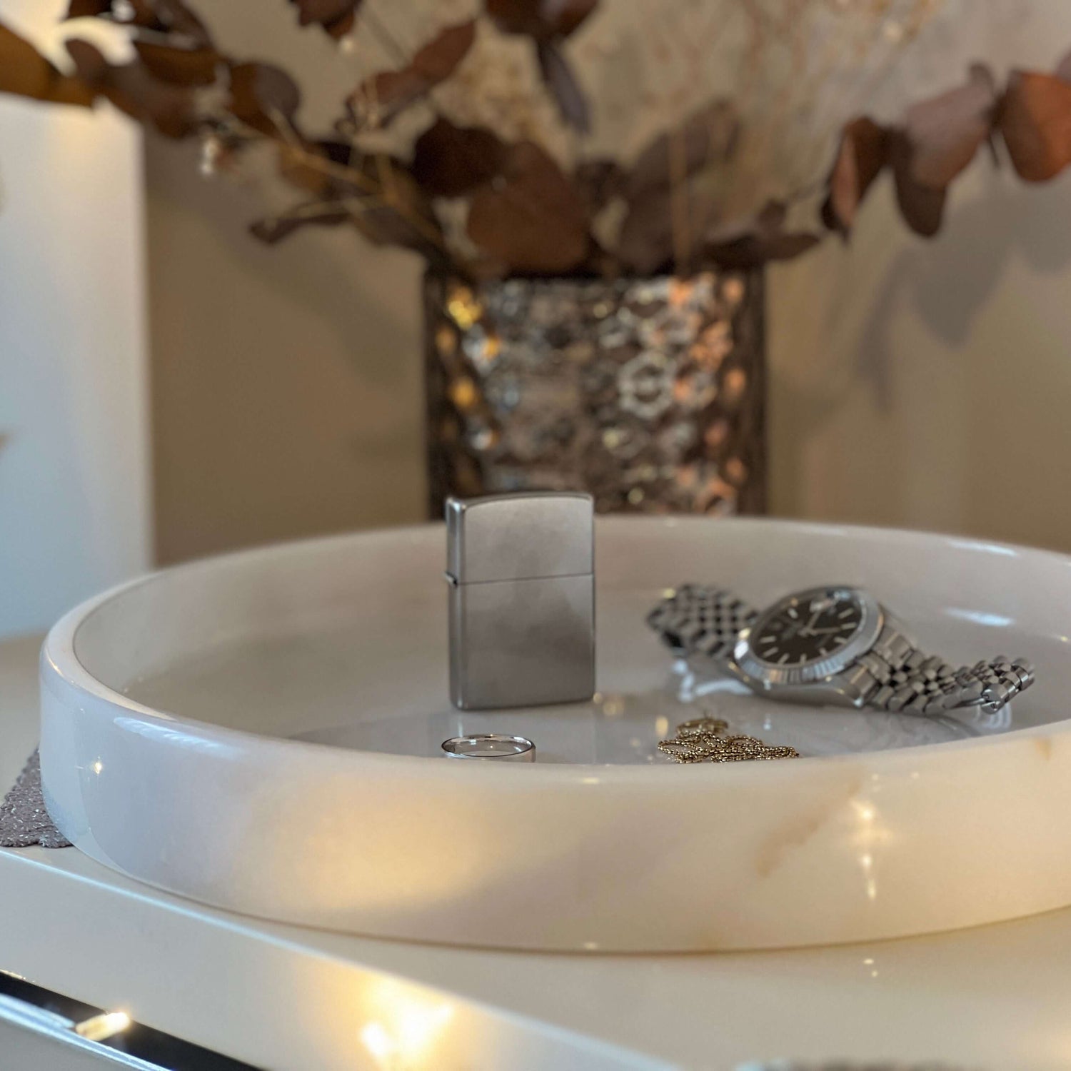 On the Travertable White Alabaster Round Tray, a silver lighter, metal band watch, ring, and gold chain are presented. Blurred dried leaves in the background enhance the cozy luxury home decor atmosphere.