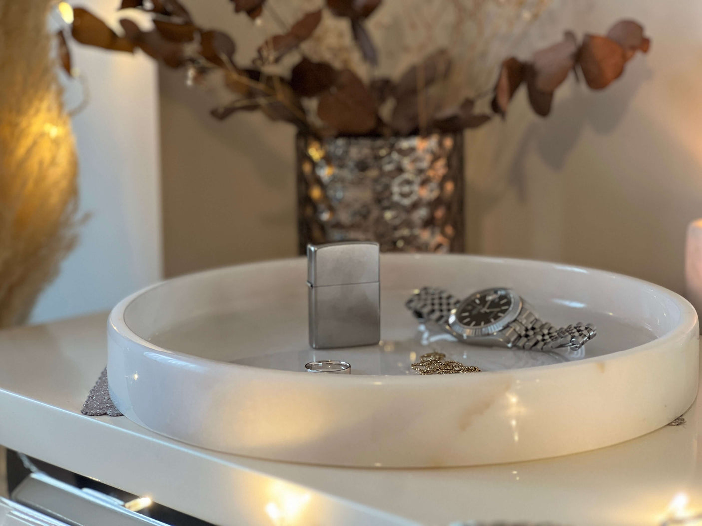 On the Travertable White Alabaster Round Tray, a silver lighter, metal band watch, ring, and gold chain are presented. Blurred dried leaves in the background enhance the cozy luxury home decor atmosphere.