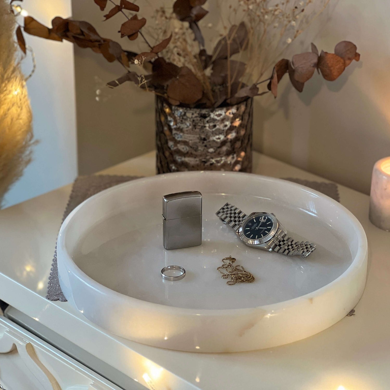 The White Alabaster Round Tray by Travertable, perfect for vanity and organizational elegance, holds a wristwatch, silver lighter, ring, and necklace. Situated on a draped white dresser under warm lighting with a lit candle and vase of dried leaves in the background, it exudes luxury home decor.