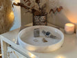 The White Alabaster Round Tray by Travertable, perfect for vanity and organizational elegance, holds a wristwatch, silver lighter, ring, and necklace. Situated on a draped white dresser under warm lighting with a lit candle and vase of dried leaves in the background, it exudes luxury home decor.
