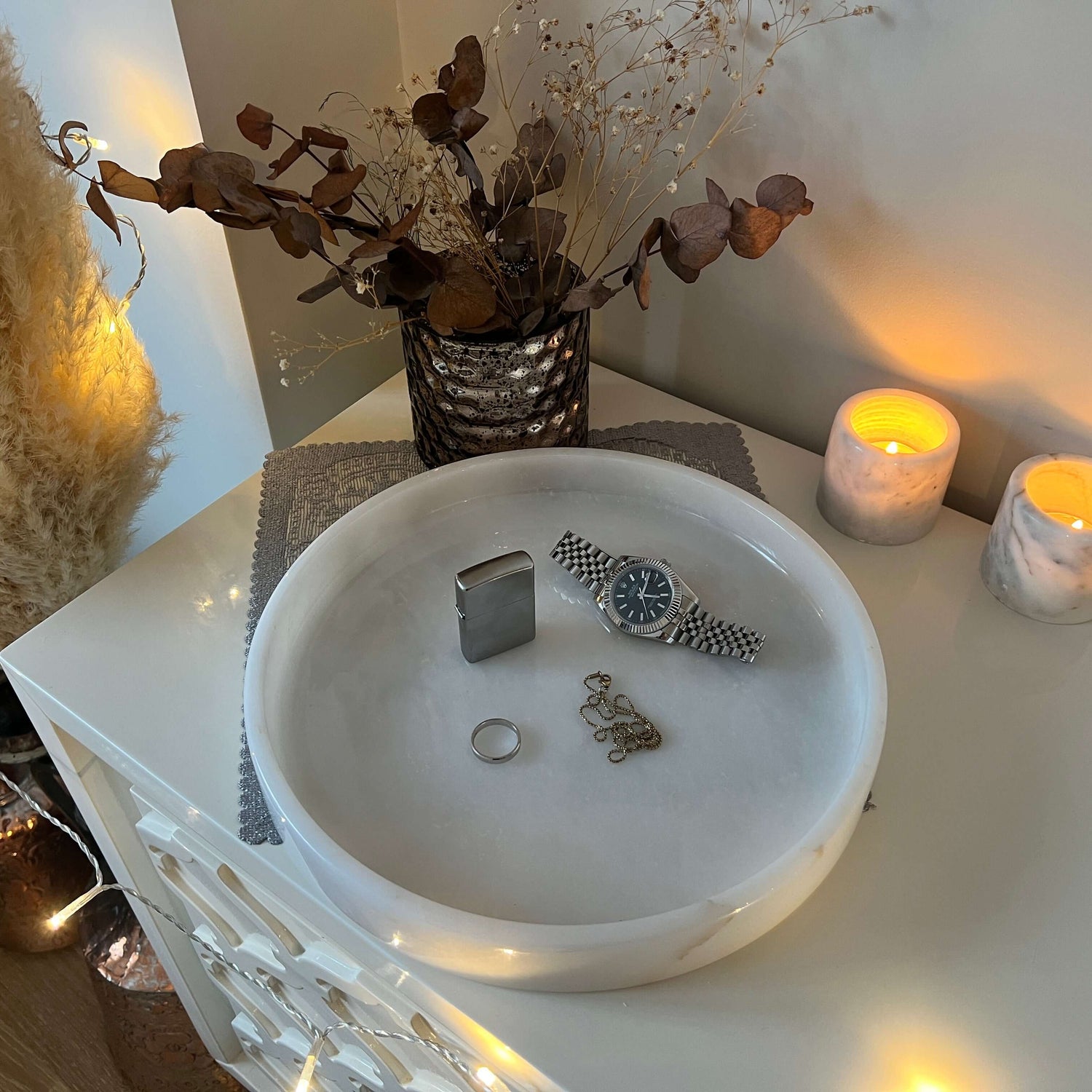 A luxury setup on a Travertable White Alabaster Round Tray showcases a silver watch, ring, and chain bracelet. Two lit candles in marble holders cast a warm glow, with dried flowers and metallic vase amid string lights and pampas grass in the background for elegant decor.