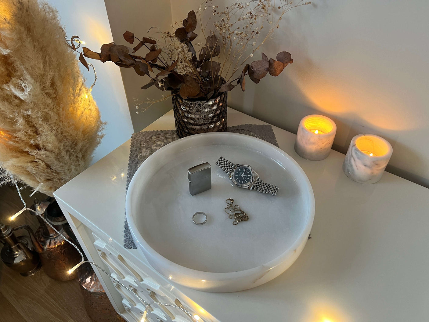 A luxury setup on a Travertable White Alabaster Round Tray showcases a silver watch, ring, and chain bracelet. Two lit candles in marble holders cast a warm glow, with dried flowers and metallic vase amid string lights and pampas grass in the background for elegant decor.