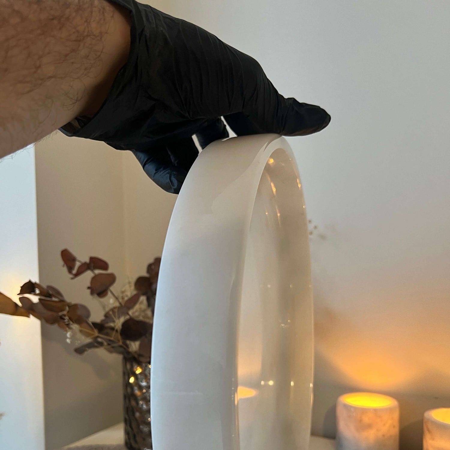 A person wearing a black glove holds the Travertable White Alabaster Round Tray vertically. In the background, lit candles, dried leaves in a vase, and a textured table runner contribute to an elegant luxury home decor scene.