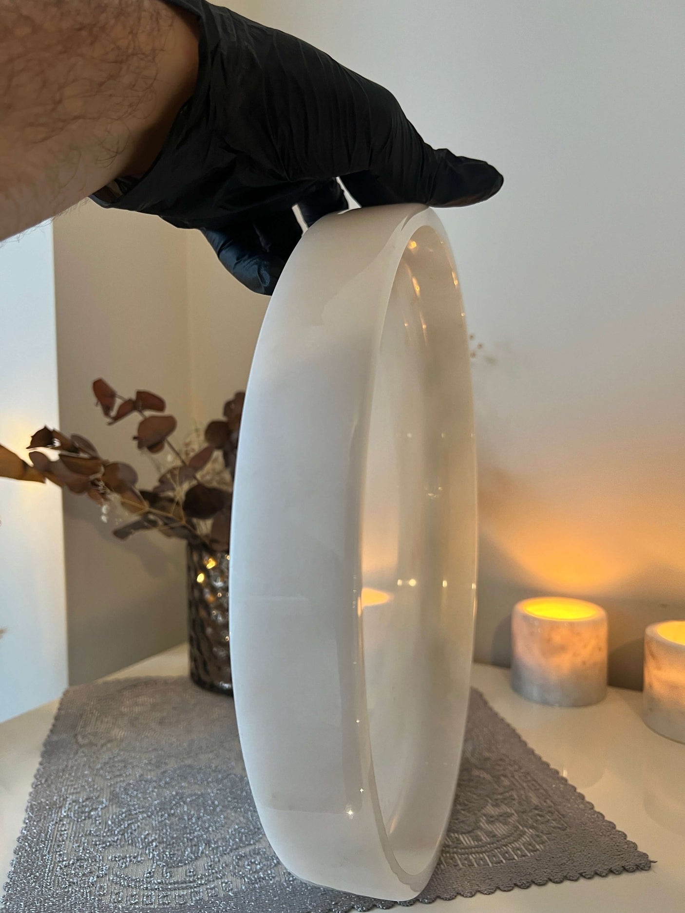 A person wearing a black glove holds the Travertable White Alabaster Round Tray vertically. In the background, lit candles, dried leaves in a vase, and a textured table runner contribute to an elegant luxury home decor scene.
