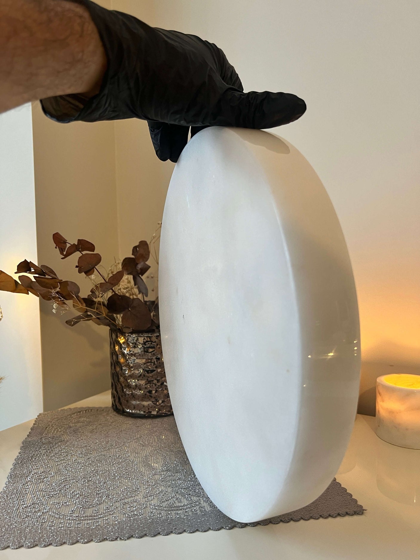 A person in a black glove holds the Travertable White Alabaster Round Tray, perfect for vanity and serving, on a tabletop. Behind, luxury decor features a decorative pot with dried brown flowers and a lit candle on a gray placemat.