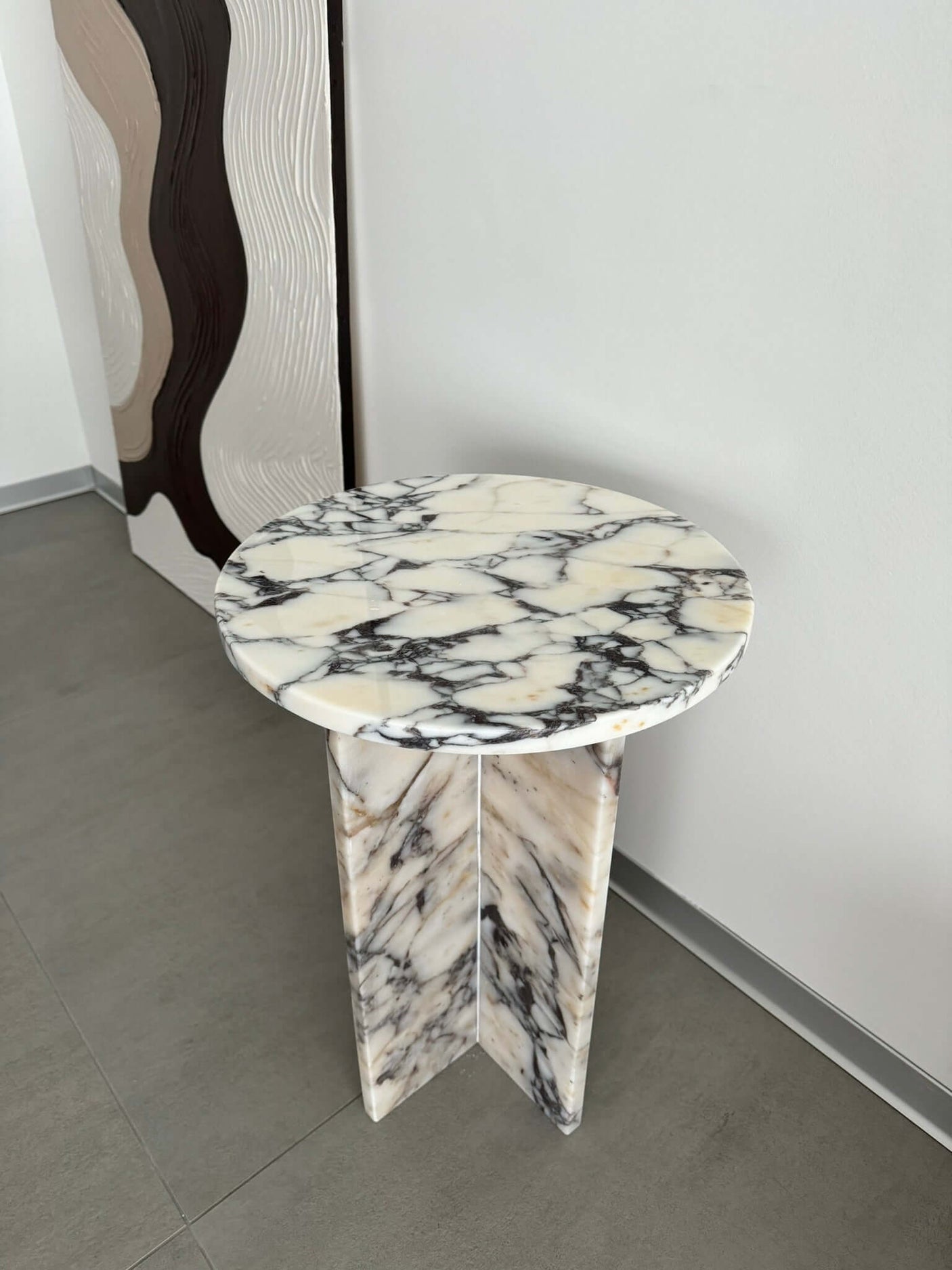 The Ela Prestige Calacatta Viola Marble Coffee Table by Travertable, a small round marble piece with a veined black and white pattern, stands on a gray floor. It features a triangular base. Behind it is a wall with textured panels displaying wavy black, brown, and beige patterns.