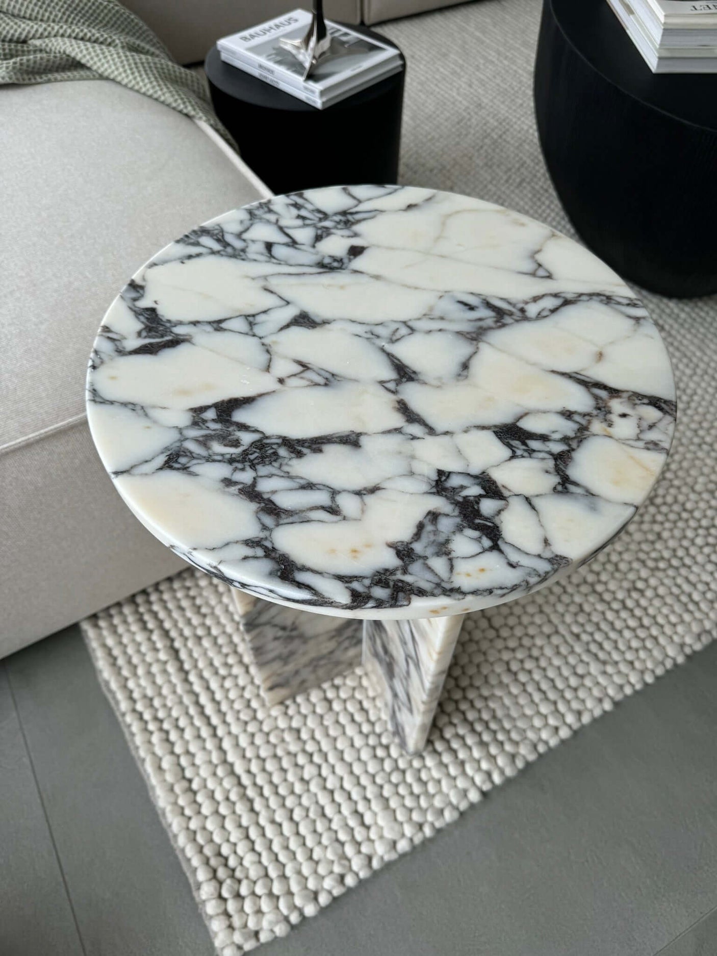 The Ela Prestige Calacatta Viola Marble Coffee Table from Travertable graces a light-colored textured rug. Nearby, a geometric black vase and stacked magazines create a stylish vignette, accentuating this handcrafted marble pieces exquisite craftsmanship and luxury.