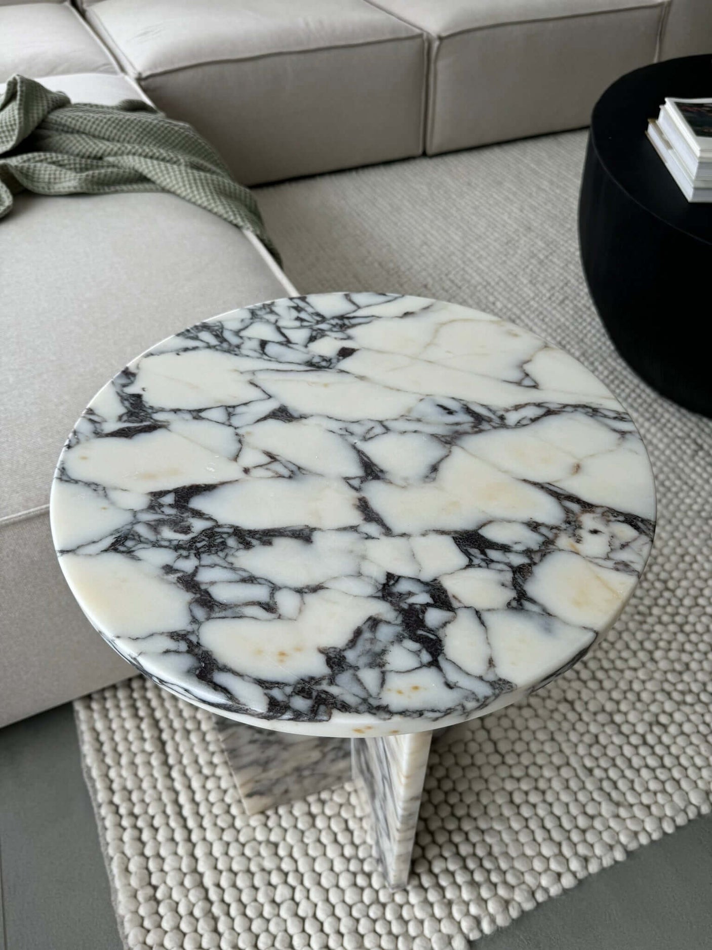 An Ela Prestige Calacatta Viola marble coffee table by Travertable, with a white and dark vein pattern, sits on a textured beige rug. Its beside a light sofa with a green blanket and a black circular coffee table holding books.