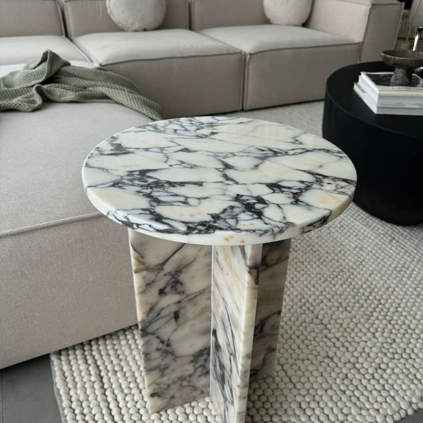 The Ela Prestige Calacatta Viola marble coffee table by Travertable, with its black and white veining, sits on a textured rug. Nearby, a light beige sectional sofa with a green throw adds harmony to the scene. The black coffee table is adorned with books and decor, enhancing the rooms charm.