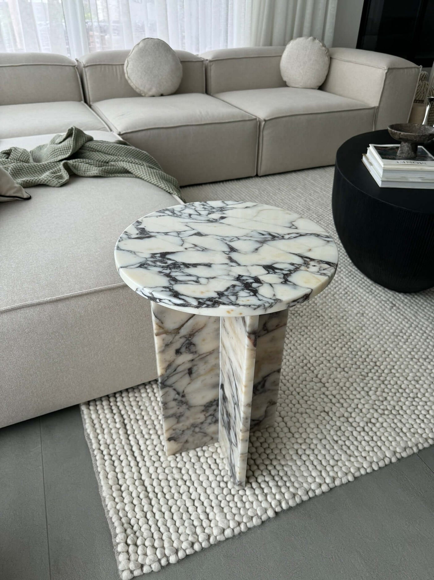 The Travertable Ela Prestige Calacatta Viola Marble Coffee Table, a luxurious handcrafted piece, sits on a textured white rug beside a modern beige sofa. A folded green throw blanket elegantly drapes over the sofas armrest, enhancing the cozy setting.