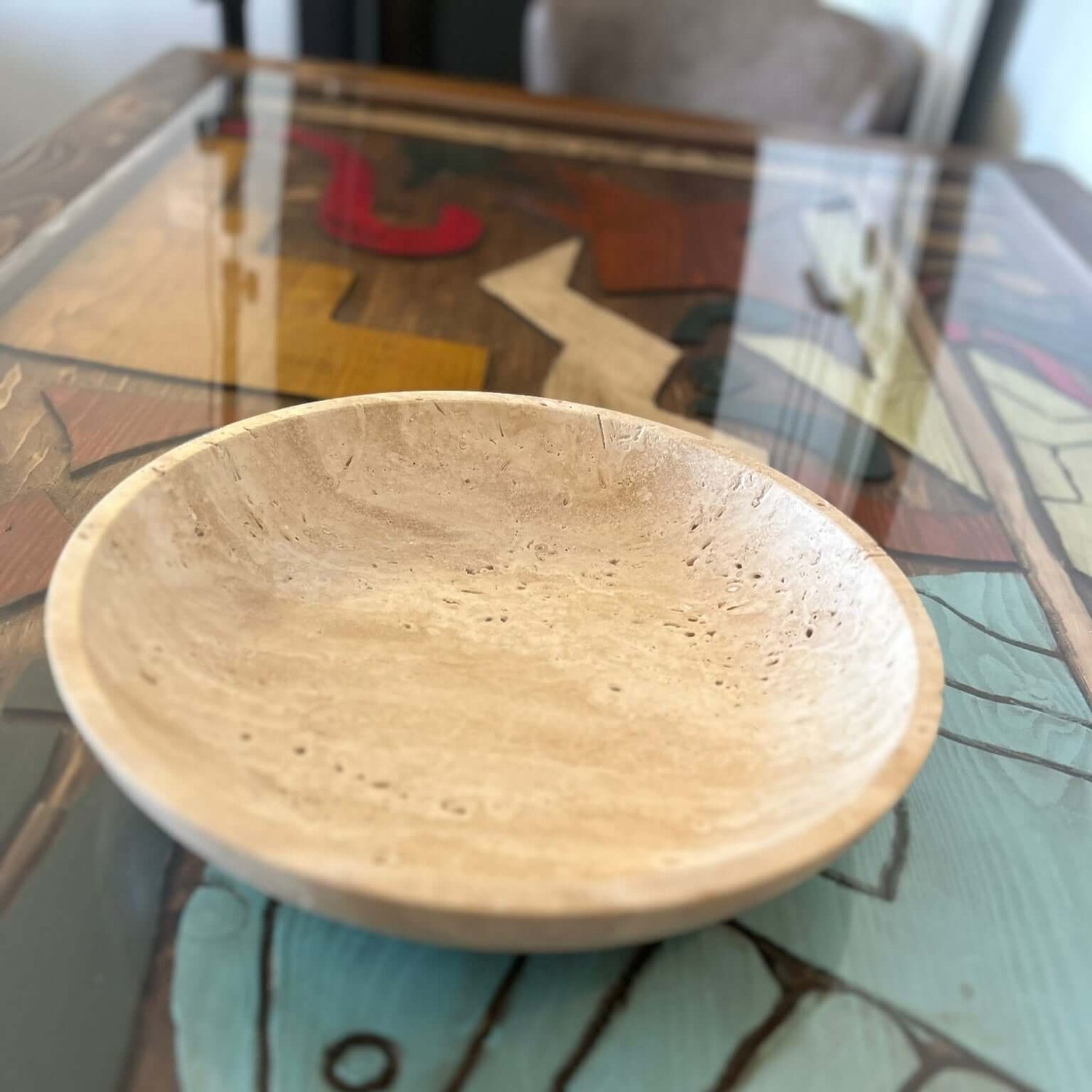 The Timeless Travertine Artisanal Bowl by Travertable, with its smooth finish and visible grain patterns, rests on a glass tabletop showcasing a colorful abstract design, bringing elegance and creativity to your space.